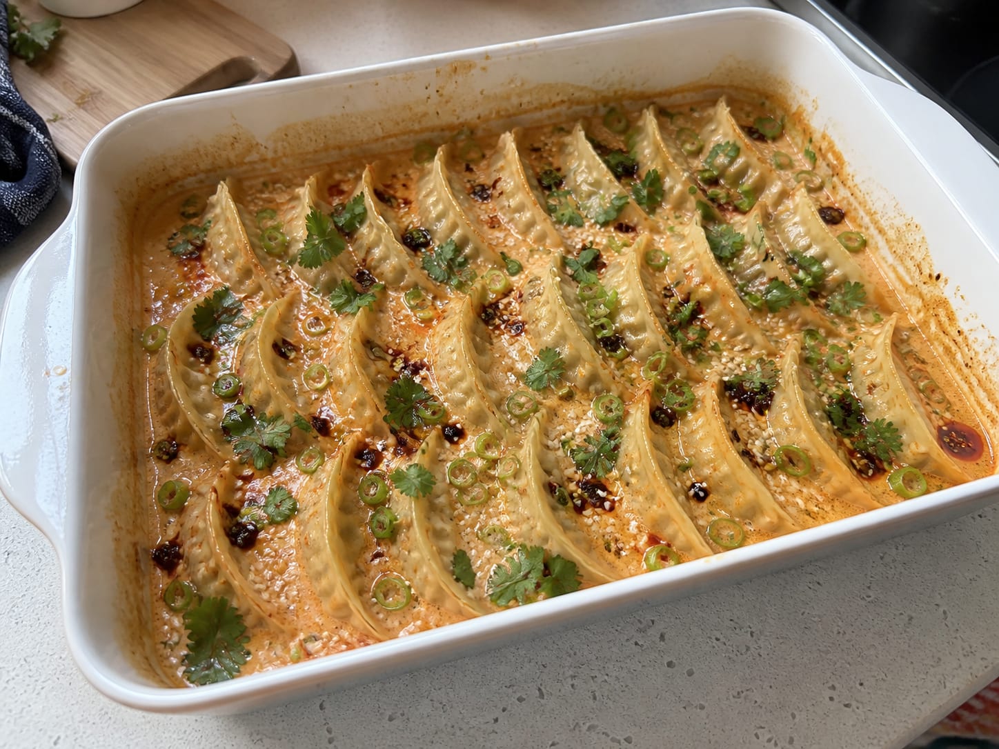 A white baking dish of dumplings baked in a creamy orange curry broth, topped with sliced spring onion, fresh coriander, sesame seeds, and crispy chilli oil
