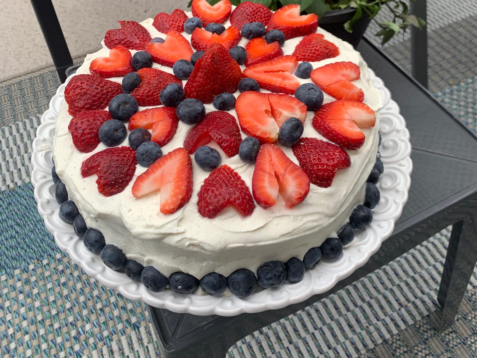 A round cake under thick vanilla-flecked cream, the top covered in halved strawberries and blueberries with a ring of blueberries around the base, on a white stand outdoors